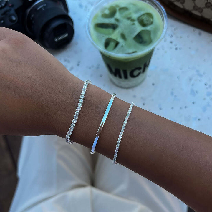 Alice Tennis Bracelet worn on wrist alongside other bracelets with a green drink in background.