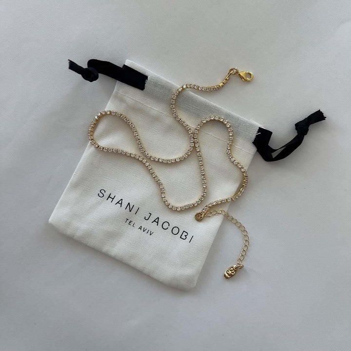 Tennis necklace by Shani Jacobi in a drawstring pouch, featuring 14K gold plated brass and crystal stones.