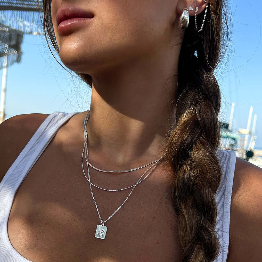 A close-up of a woman wearing a Sunset Necklace and silver earrings against a sunny outdoor backdrop.